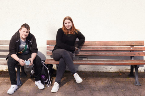 Schulhof: 2 Schüler:innen sitzen in der Sonne auf einer Bank